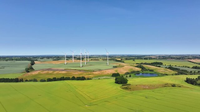 Aerial view of wind turbines standing in vast green and yellow fields with a small pond under a clear blue sky in Uckermark, Brandenburg, Germany.