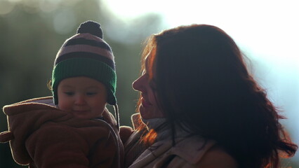 Mother kissing baby on cheek while child wears bear teddy fleece coat and striped beanie hat outdoors, parent showing affection and love during family bonding in nature © Marco