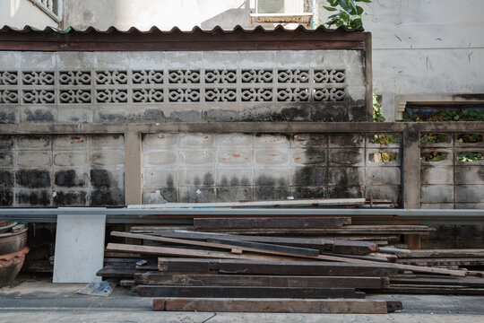 A pile of weathered wooden boards and scrap building materials lies on the ground in front of an old, heavily stained concrete block wall featuring decorative breeze blocks.