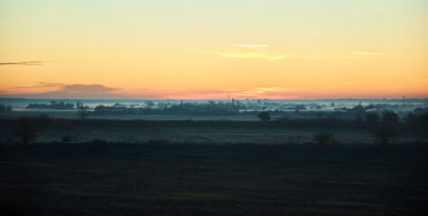 Obraz premium A misty dawn envelops the rolling fields and distant trees. The twilight sky is painted in soft pink and orange tones. Fog envelops the grass