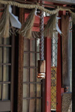 Thick braided rope hangs from Suzu bell in small inner-city shrine partially illuminated by sunlight filtering through nearby buildings, capturing still moment of Japanese tradition and reverence.