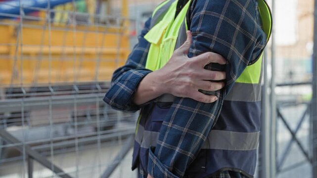 Man builder clutching upper arm and gripping forearm at building site wearing reflective vest and scaffolding behind; injury pain.