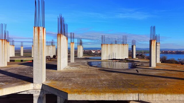 Rooftop view of building under development featuring concrete pillars with steel armature and water puddles. Top floor of concrete construction site with vertical rebar. Aerial view.