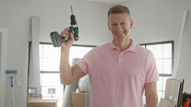 Man in pink polo holds a cordless drill aloft with raised hand and visible forearm inside a bright residential building room; confidence home improvement progress.