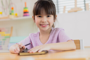 Naklejka premium Cute successful learner of primary school, smiling asian School girl sitting in school writing in book with pencil, studying, education, learning. Asian children in the class. Student diversity.