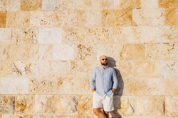Obraz premium Bald man wearing sunglasses and casual summer clothes standing relaxed against a textured stone wall under warm sunlight