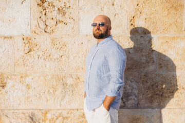 Obraz premium Bald man with beard and sunglasses standing by stone wall in Cagliari, Sardinia enjoying sunshine