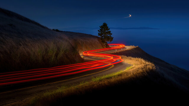 Long exposure light trails on a winding mountain road at night, glowing red car tail lights on a curved coastal highway, dark blue twilight landscape