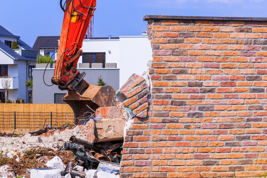 Construction equipment breaks down a brick wall during demolition work in a residential area on a sunny day