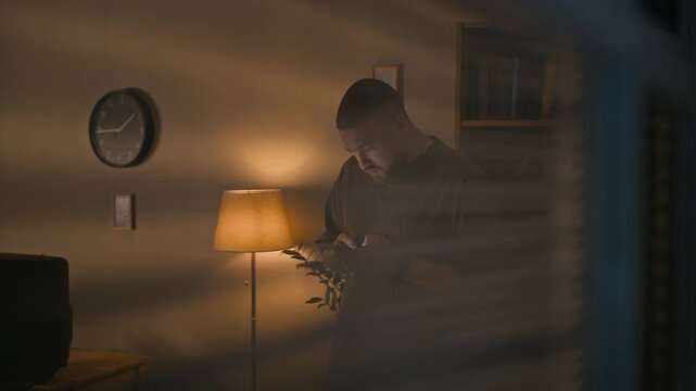 Through window blinds view of bearded Caucasian man texting on smartphone in dimly lit apartment illuminated by warm lamp light