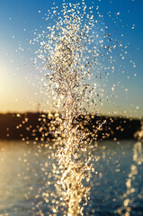 Fototapeta premium Splashes of water from fountain at sunset against pond. Golden reflections. Atmospheric evening view