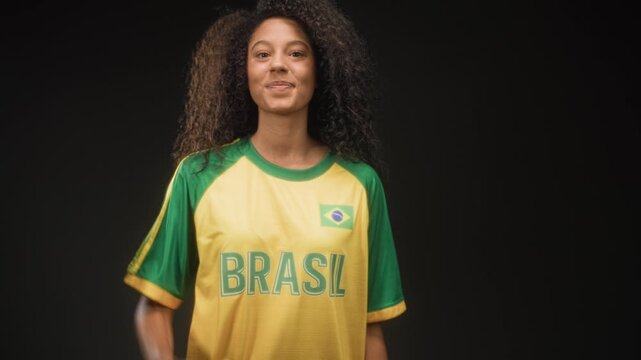 Woman points finger to camera with a smiling face, wearing yellow green brasil football tshirt with flag patch in studio; joyful support.