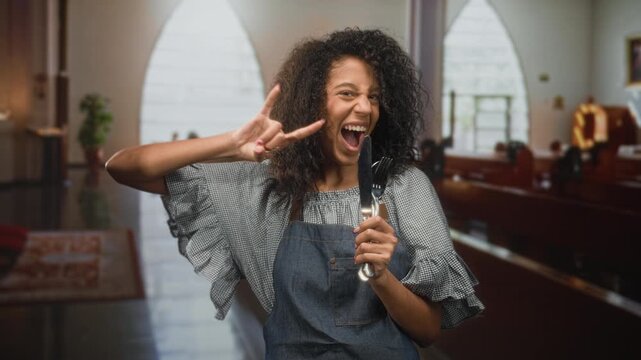 Woman in apron holding fork and knife, mouth open and right hand peace sign in building; playful celebration.