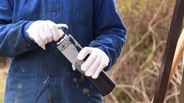 Woman in blue overalls preparing to paint outdoors. Construction worker holding a bottle of black paint, close-up. Worker in blue overalls and white protective gloves holding a plastic can of paint