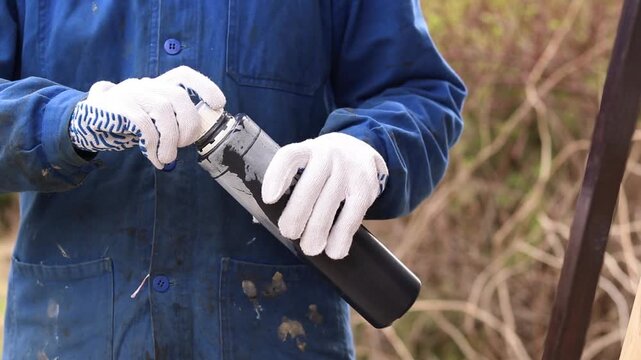 Woman in blue overalls preparing to paint outdoors. Construction worker holding a bottle of black paint, close-up. Worker in blue overalls and white protective gloves holding a plastic can of paint
