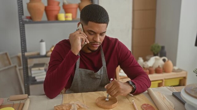 Hispanic man artist with beard shaping a clay bowl using a sculpting tool while holding smartphone to his ear at a pottery studio surrounded by pots and tools; concentration.