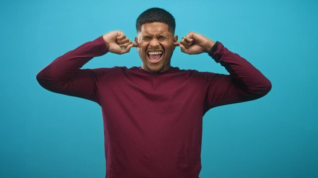 Hispanic man with beard plugs ears with fingers while grimacing in studio; frustration sensory overload.