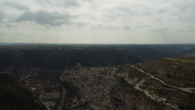 4K aerial drone descent, revealing the dramatic Alcal&aacute; del J&uacute;car village built into a cliff side within a canyon, with the vast Castile-La Mancha plateau in the background