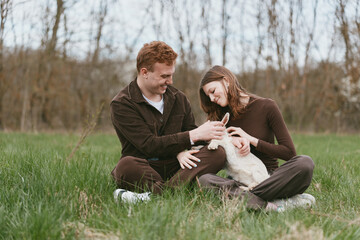 Naklejka premium A smiling couple sits on the grass in a park, cradling a young goat kid between them. A candid lifestyle moment highlighting connection, nature, and playful companionship.