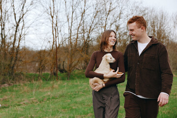 Naklejka premium Happy couple outdoors with a small white dog, smiling at each other, embracing in a green field, casual denim and sweaters, candid lifestyle moment. Natural light enhances smiles and warmth.