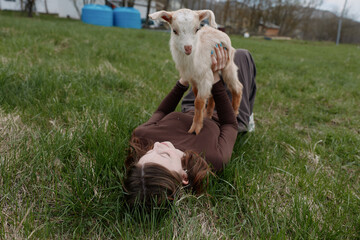 Naklejka premium Young woman lies on grass holding a small goat above her with a joyful expression in a natural outdoor setting during a casual lifestyle moment.