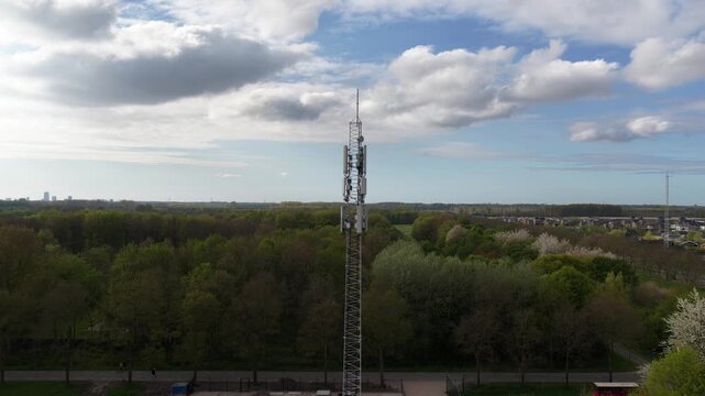 Descending aerial shot alongside a tall telecommunication tower revealing its lattice steel structure, panels, and antenna against a backdrop of a lush green forest and a partly cloudy sky