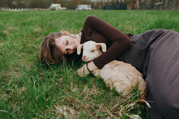 Naklejka premium Joyful woman outdoors, lying on grass, cuddling a goat kid, sunlit moment, relaxed posture, gentle breeze, natural colors, pastoral setting, candid lifestyle in a peaceful countryside field.