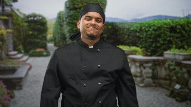 Man chef in uniform points finger to camera with right hand and places fist to chest in a building courtyard; pride culinary craft.