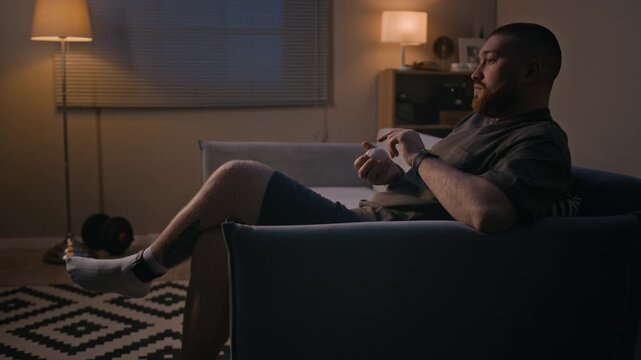 Side view of Caucasian man under house arrest sitting on couch with baseball while looking toward window with flashing red and blue lights in apartment at night