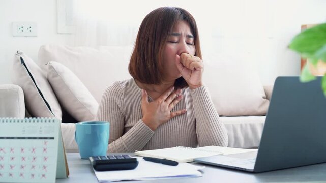 Sick Asian woman coughing and holding chest while working from home on laptop. Concept of illness, cold, flu, and remote work health issues during winter