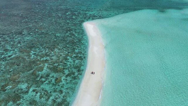 Aerial view of a narrow white sandbar with two people walking on it, surrounded by crystal clear turquoise water and coral reefs in Thoddoo, Alif Alif Atoll, Maldives.