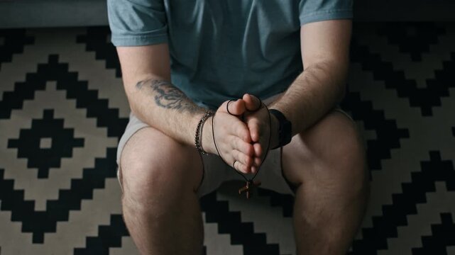 From above tilt up shot of man wearing electronic ankle bracelet sitting on floor in apartment and praying with cross in hands during difficult home confinement period