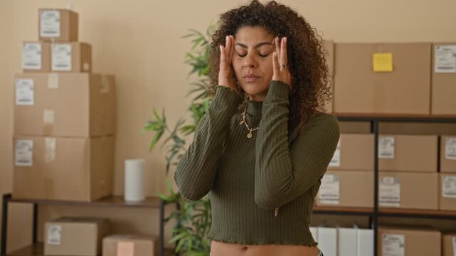 Woman pressing temples with hands amid stacked shipping boxes and parcels in a studio packing area for a small business, eyes closed and tense; stress.