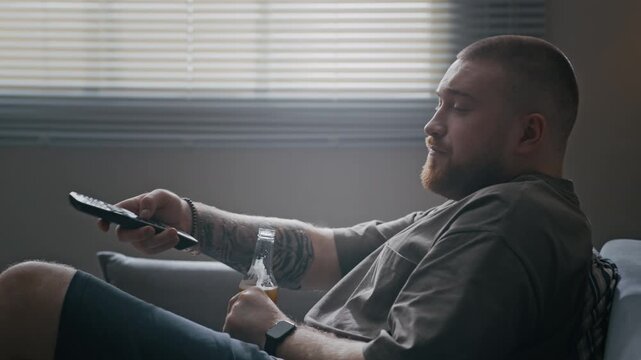 Tilt down of Caucasian man drinking beer on couch while watching TV in apartment and spending quiet time alone during home restriction