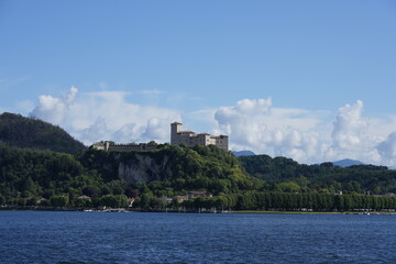 Naklejka premium View of the historic Rocca di Angera fortress rising above the shores of Lake Maggiore in Lombardy, Italy. The medieval castle stands on a green hillside under a clear blue sky.