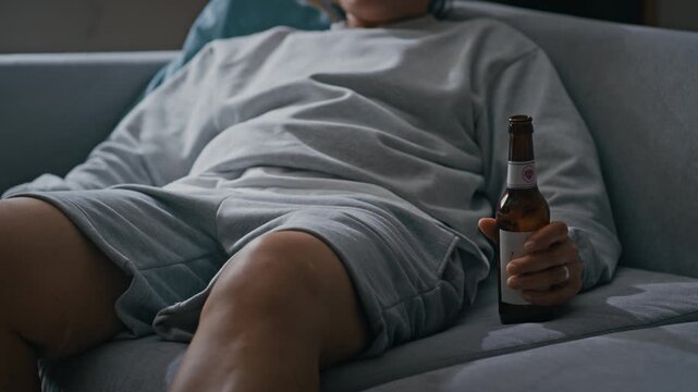 Tilt down shot of Biracial woman with electronic ankle bracelet drinking beer while watching TV alone at home in apartment living room