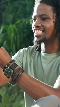 Vertical video: Checking watch, man with locs in green tee grabbing tablet on grass, checking cable
