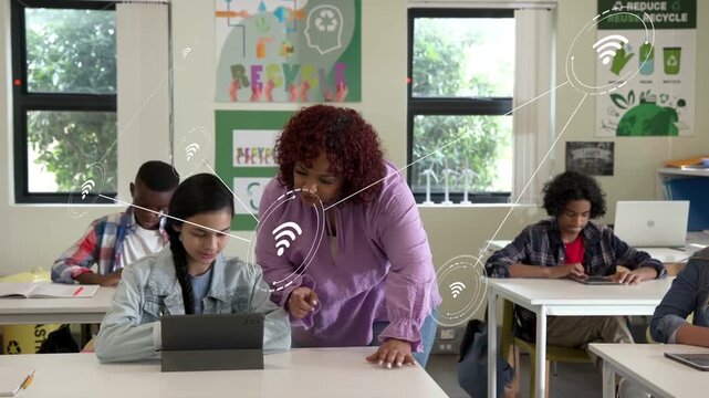 Teacher leaning over pupil tapping tablet, pointing directing class as wifi icons linking to tablet