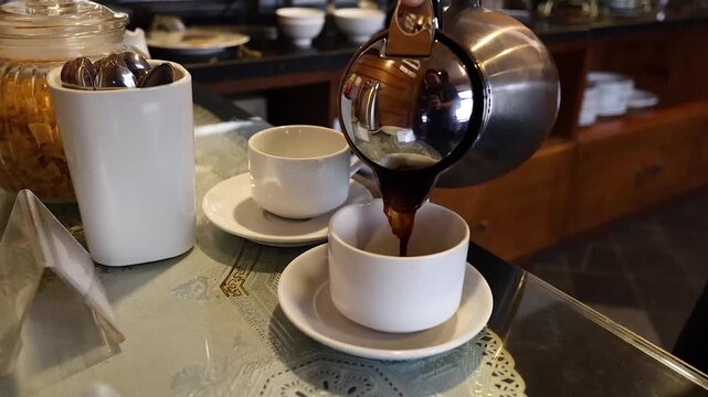 Close up motion of person pouring fresh hot black coffee from metal pot into white ceramic cups on table.