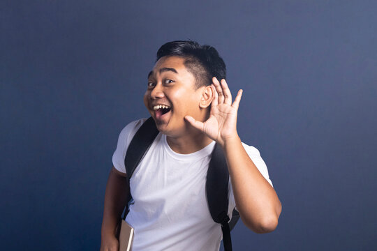 Young man wearing a backpack holding a hand to his ear as if listening or eavesdropping with a happy and excited facial expression.