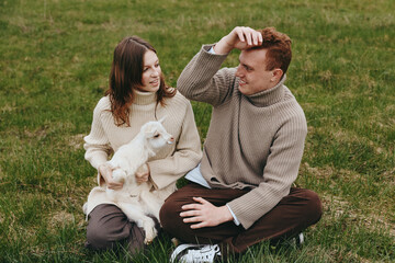Naklejka premium Couple sitting on grass with a white goat kid, wearing sweaters, enjoying a relaxed outdoor lifestyle moment with nature, affection, and gentle companionship in warm lighting, cozy ambiance.