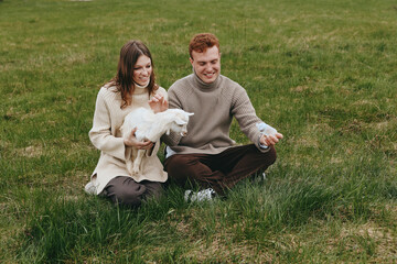 Naklejka premium Couple sitting on grass with a playful goat kid, sharing a gentle moment outdoors. Cozy sweaters, natural light, warm countryside mood. They interact tenderly, suggesting lifestyle, wellness, and