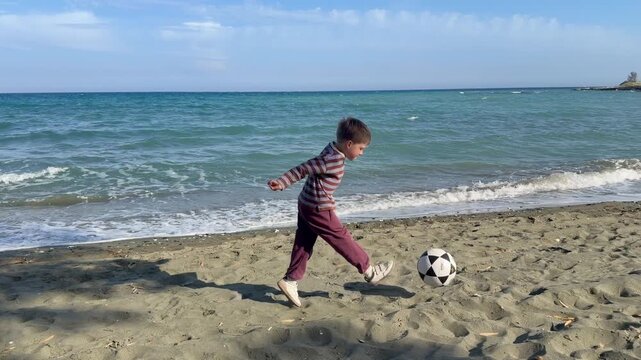 Child plays soccer on sandy beach shore with sea waves and blue sky in background during summer vacation. Active outdoor play footage suits family resort promotion, kids sportswear, vacation content