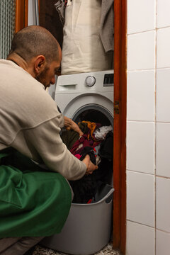 Hispanic bearded man in green apron taking clean laundry from washing machine