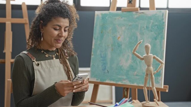 Woman scrolls smartphone with hands in art studio while wearing apron and viewing canvas and wooden mannequin pose; calm inspiration.