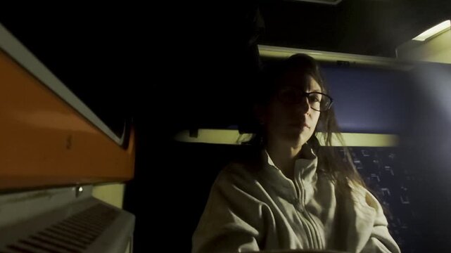 A professional static shot captures a traveler in glasses seated in the shadows of a quiet train compartment at night. Soft ambient light from above creates a somber and reflective atmosphere.