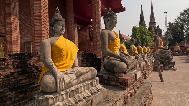 A cinematic motion footage showing rows of ancient stone buddha statues adorned with yellow sashes under a clear sky.