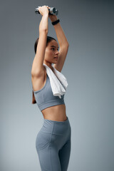 Obraz premium Female fitness enthusiast lifts a dumbbell overhead with a towel around the neck, wearing athletic gear in a studio setting, showcasing strength, focus and training.