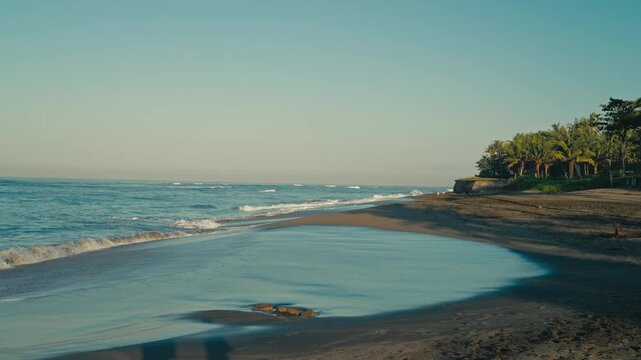 Erosion patterns on a black sand beach fringed by palm trees slowly disappear into the ocean as calm waves wash ashore. Minimal tropical scene showing natural change and coastal textures.