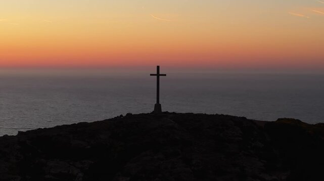 A lone cross stands on a hilltop, silhouetted against a soft dawn sky with the calm sea stretching beyond. Ideal for spiritual themes or coastal landscape features.